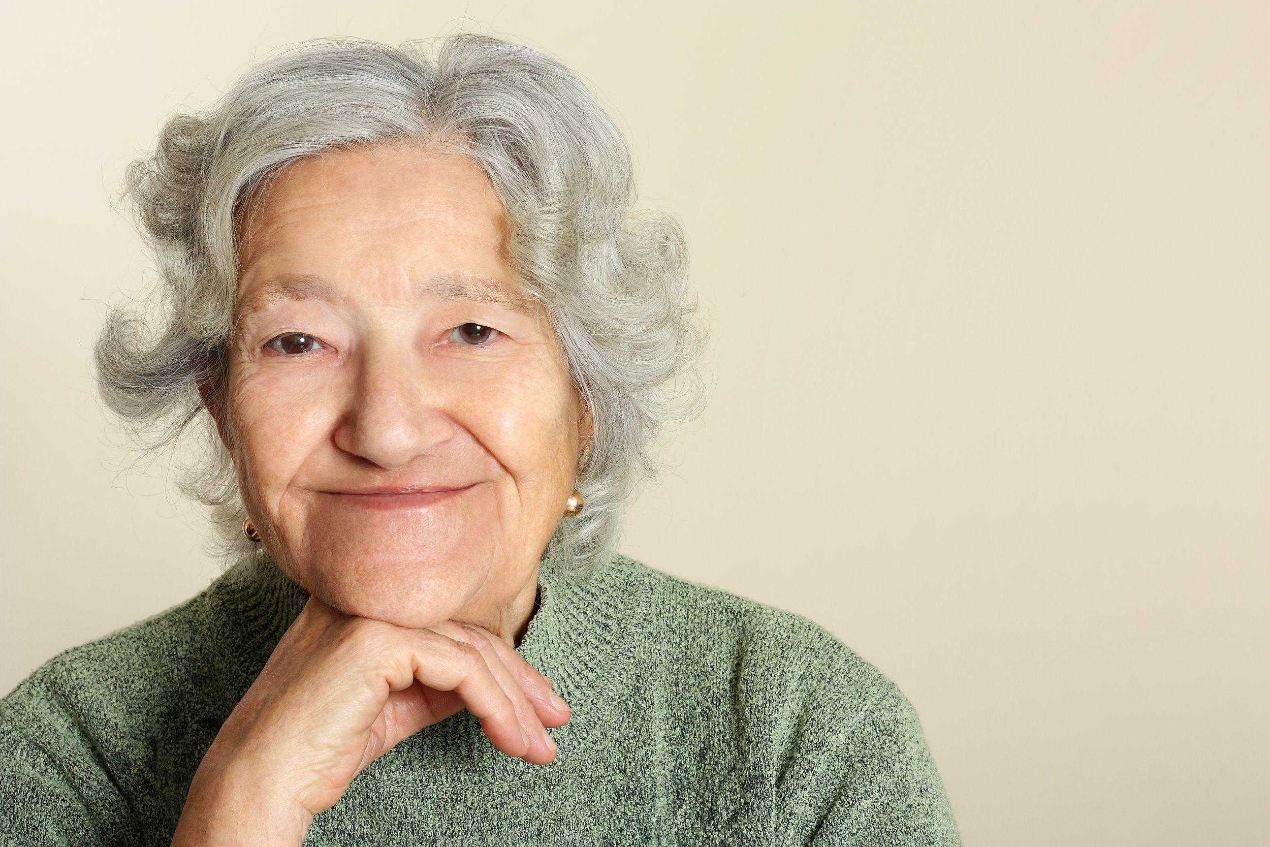 Elderly woman with gray hair smiling and resting her chin on her hand, wearing a green sweater, with a neutral background.