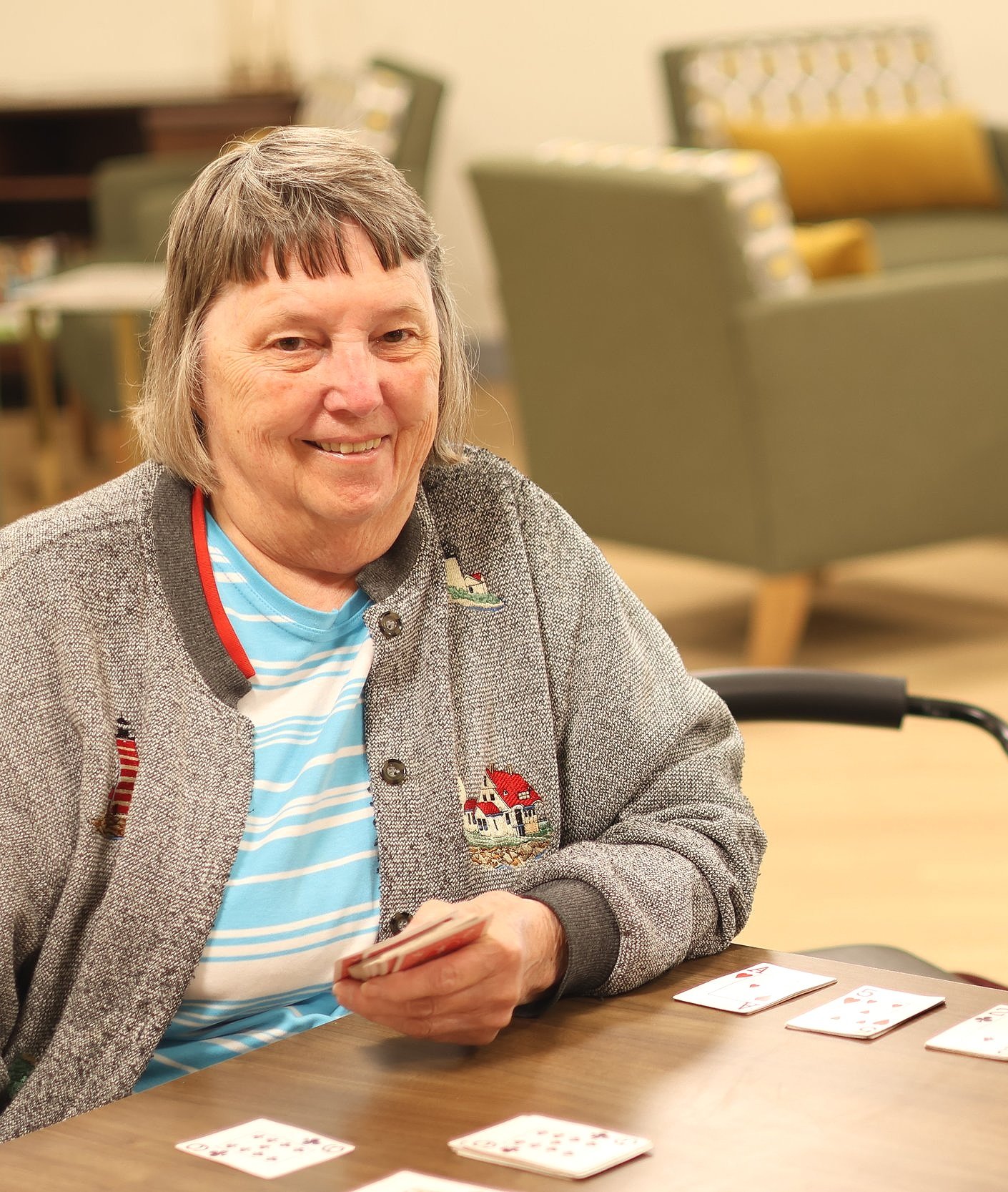 Elderly woman playing a card game, seated at a table with playing cards spread out, in a cozy room with armchairs in the background.