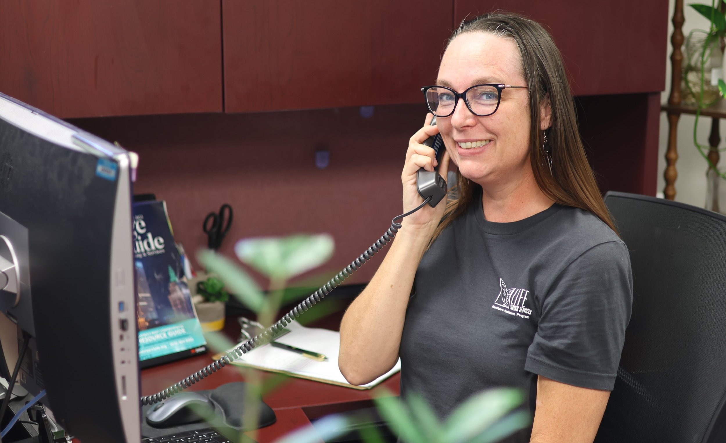 Woman smiling while talking on a desk phone, sitting at an office desk with a computer monitor and office supplies.