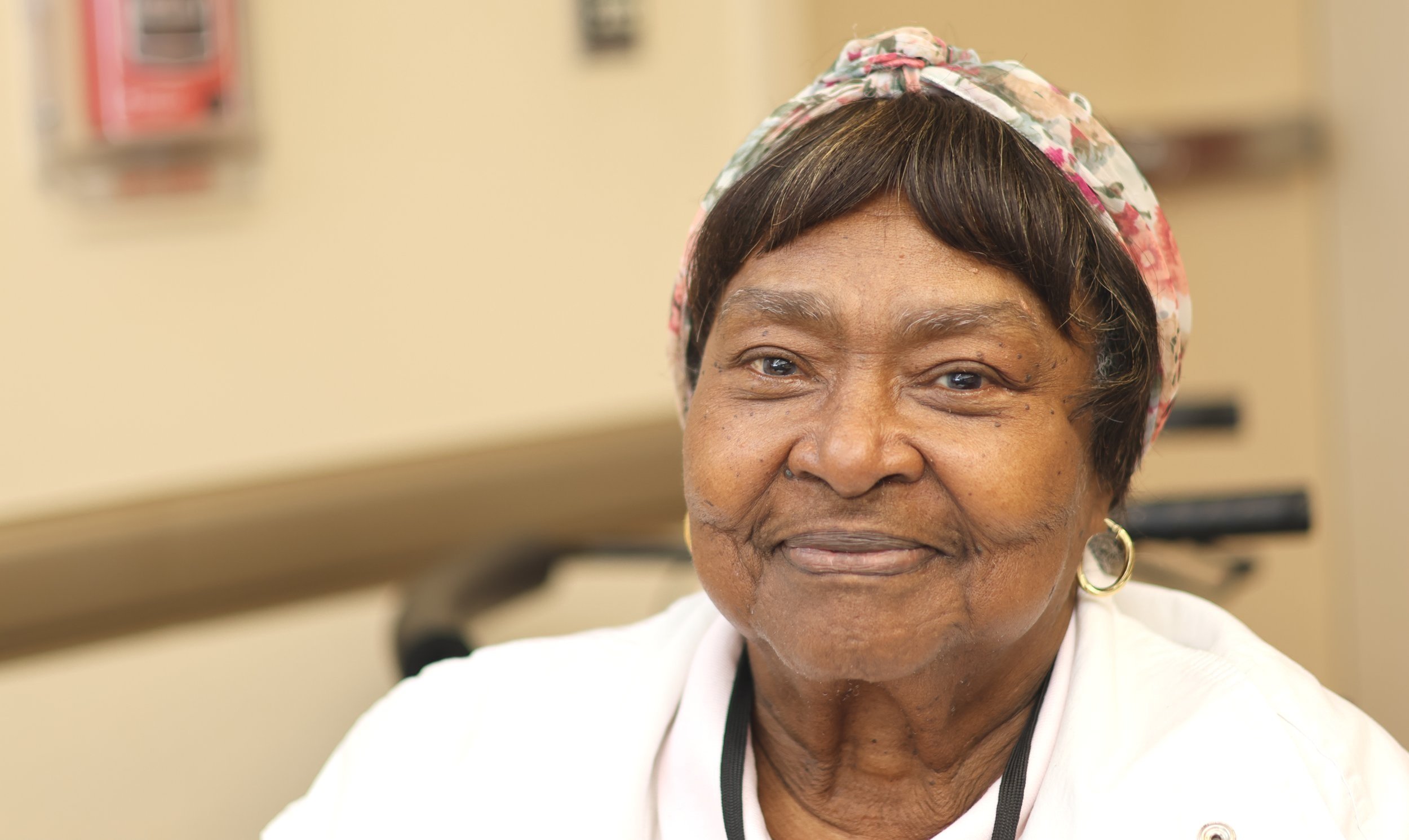 Elderly woman smiling with a floral headscarf and white clothing.