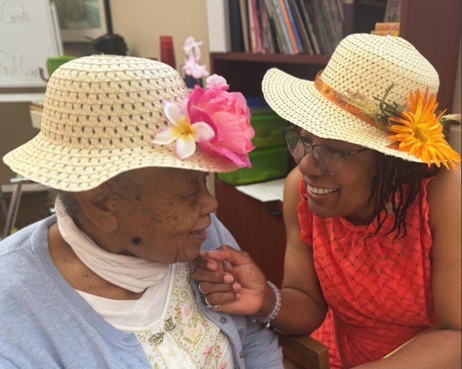 Two women wearing straw hats with floral decorations, smiling and talking to each other indoors.