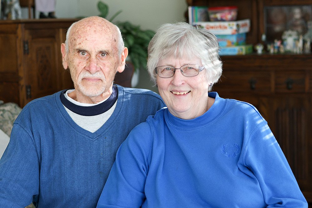 Elderly couple smiling indoors with wooden furniture in the background.