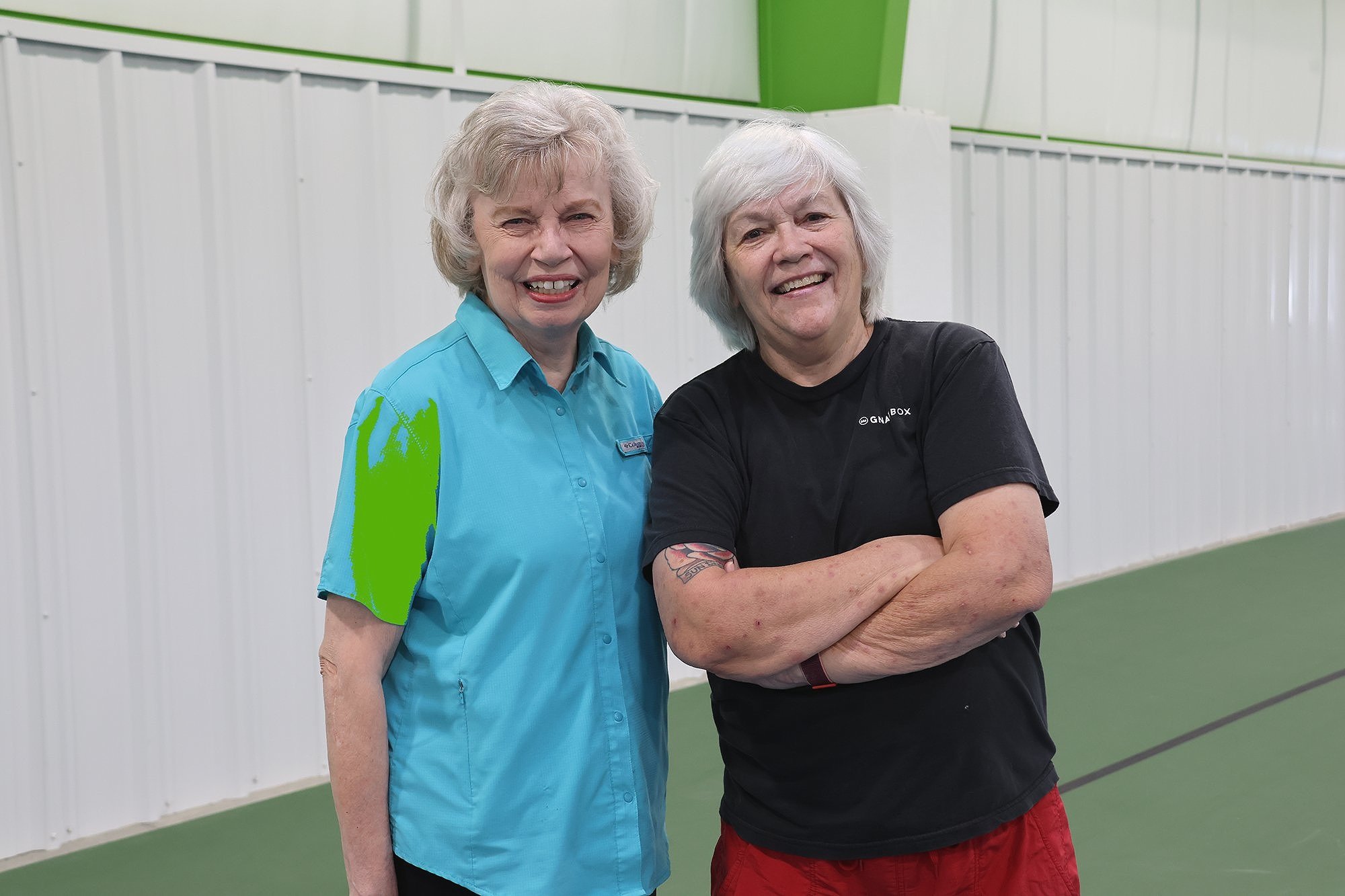 Two older women smiling and standing in a sports facility, with one wearing a blue shirt and the other a black T-shirt and red pants.