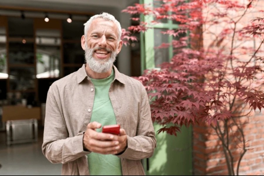 Older man with gray hair and beard smiling while holding a red smartphone outdoors, standing next to a red maple tree.